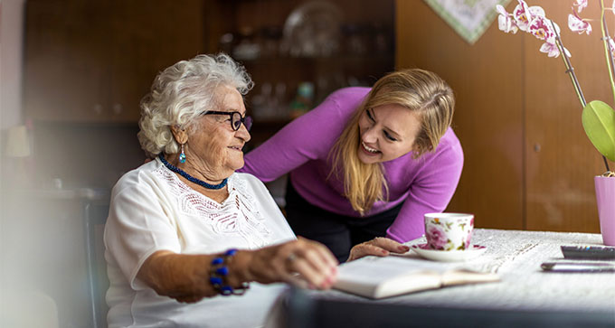 a female carer in a pink cardigan smiling at an elderly lady sitting down in a chair at a table