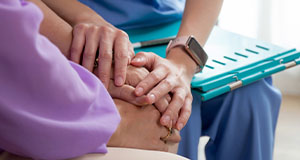 a nurse in blue with a clipboard holding the hands of a purple-clothed elderly person