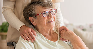 an elderly woman sitting in a wheelchair holding the hand of a carer behind her