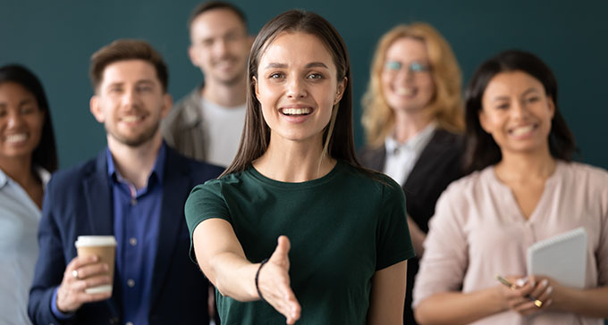 a group of smiling people in smart clothing, with the woman in front extending her hand to the viewer