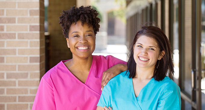 two nurses in pink and blue scrubs outside and smiling ahead