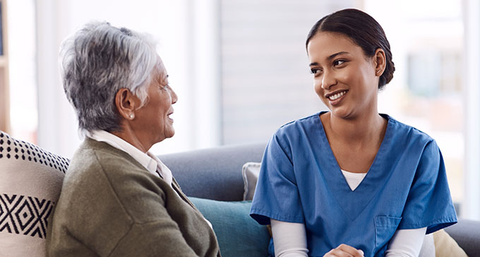 a nurse sitting on the sofa next to an elderly woman conversing with one another