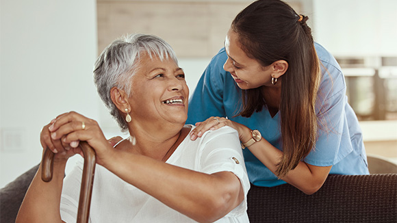 an elderly lady with a cane sitting on a sofa and smiling as a carer who is leaning from behind the sofa